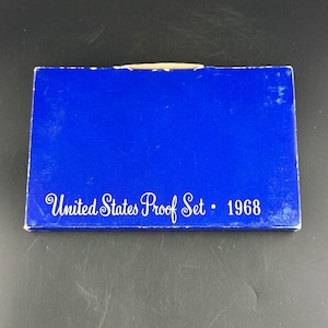 May include: A white ceramic mug with a black handle and the text "But First, Coffee" in a black, bold font. The mug is on a white surface.