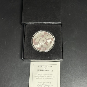 May include: A silver coin featuring a panda bear design, presented in a black box with a certificate of authenticity. The coin has the text "10元" and "1oz Ag .999". The certificate is signed by Nicholas J. Bruyer, Director, Preferred Customer Club.