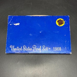 May include: A blue box with the text "United States Proof Set • 1968" in white script. The box has a yellow circle with a brown design on the top right corner. The box is slightly worn.