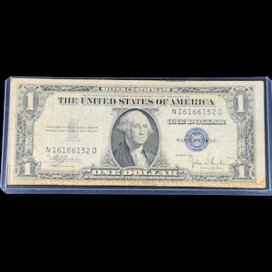 May include: A one-dollar silver certificate from the United States of America. The bill features a portrait of George Washington and the text "ONE DOLLAR" in multiple locations. The bill is encased in a clear protective sleeve.