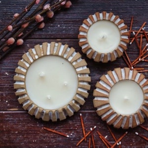 Op de afbeelding: Drie decoratieve kaarsen in keramische houders met een beige en wit ontwerp. De kaarsen zijn gevuld met witte was en hebben meerdere lonten. Lucifers en takken met roze knoppen zijn rond de kaarsen verspreid.