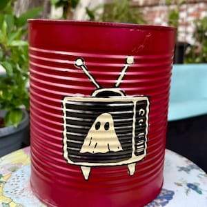 May include: A cylindrical red metal container with horizontal ridges. The front features a black and cream-colored graphic of a vintage television set with two antennas, displaying a simple ghost figure on the screen.