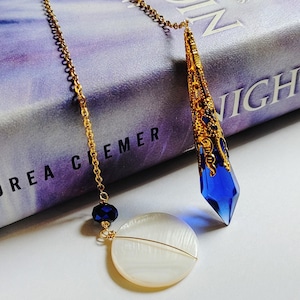 May include: A gold chain bookmark with a blue crystal pendant and a white shell charm. The bookmark is resting on a book titled "Nightshade" by  S.A. Chakraborty.