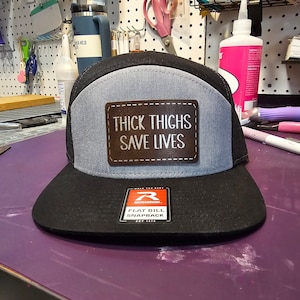May include: A black and gray flat bill snapback hat with a mesh back. The front panel is gray with a brown leather patch that reads "THICK THIGHS SAVE LIVES" in white text. The hat has a black bill and a Richardson tag.