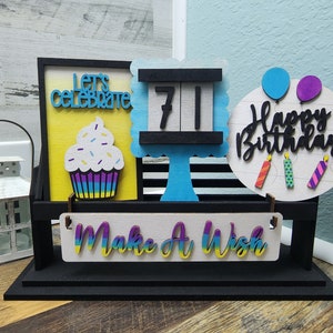 May include: A black wooden stand with a cupcake, a sign that says "Let's Celebrate", a sign that says "Happy Birthday" with balloons and candles, a sign that says "Make a Wish", and a sign with the number "71" on it.