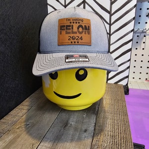 May include: A gray and black trucker hat with a brown leather patch that says "I'm voting FELON 2024" with stars above and below the text. The patch has a design that resembles the American flag.