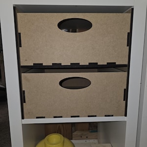 May include: Two white wooden storage boxes with oval cutouts on a white shelf. The boxes are made of light brown wood and have black trim. The shelf is part of a white bookcase.