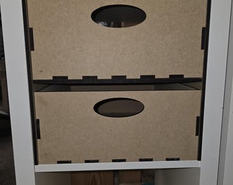 Op de afbeelding: Twee witte houten opbergdozen met ovale uitsparingen op een wit rek. De dozen zijn gemaakt van lichtbruin hout en hebben zwarte afwerking. Het rek maakt deel uit van een witte boekenkast.