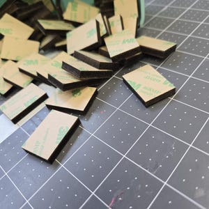 May include: A pile of black and brown adhesive squares with green 3M branding. The squares are scattered on a gray surface with a grid pattern.