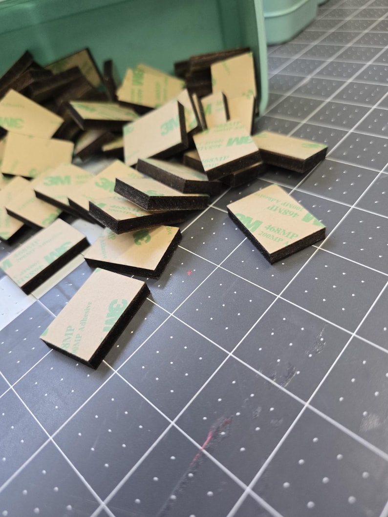 May include: A pile of black and brown adhesive squares with green 3M branding. The squares are scattered on a gray surface with a grid pattern.