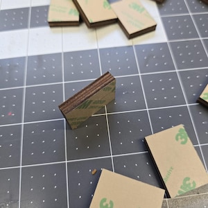 May include: A stack of small, rectangular, brown wood pieces with adhesive backing. The adhesive is labeled "3M" and "adhesive".