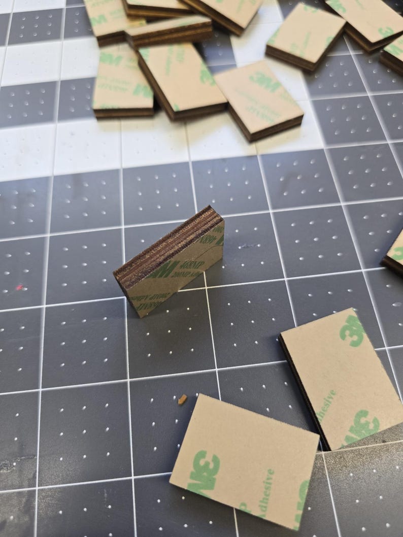 May include: A stack of small, rectangular, brown wood pieces with adhesive backing. The adhesive is labeled "3M" and "adhesive".