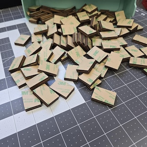 May include: A large pile of brown and green adhesive squares with the 3M logo printed on them. The squares are stacked on top of each other and are spilling out of a light blue plastic container.