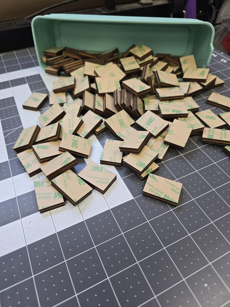 May include: A large pile of brown and green adhesive squares with the 3M logo printed on them. The squares are stacked on top of each other and are spilling out of a light blue plastic container.