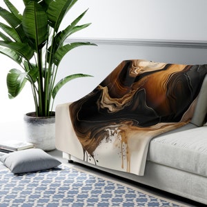May include: A soft, brown, black, and gold abstract throw blanket with a flowing, liquid-like design. The blanket is draped over a white couch.