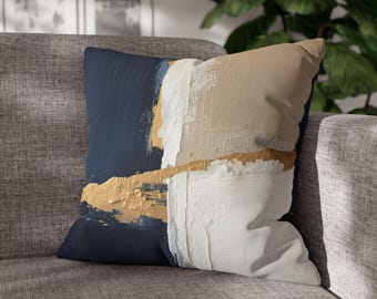 Può includere: Cuscino decorativo quadrato con un design astratto. Il cuscino presenta una sezione blu navy, una sezione bianca e una sezione beige, con accenti dorati. Il cuscino è posto su un divano grigio.