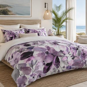 May include: A bedding set featuring a duvet cover and matching pillow shams with a watercolor-style floral pattern in shades of purple, lavender, and white. The bedding is layered over white sheets and includes a solid white top section on the duvet.