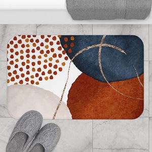 Puede incluir: Alfombra de baño abstracta con fondo blanco y formas coloridas en tonos naranja, marrón, azul y blanco. Las formas están delineadas con purpurina dorada.