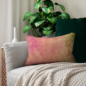 May include: A soft, light pink and yellow throw pillow with a watercolor-like design. The pillow is on a light gray couch with a white throw blanket.
