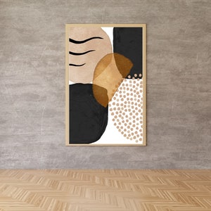 May include: Abstract art print in a light wood frame. The artwork features a composition of black, tan, and gold shapes, including a dotted pattern and curved lines. The art is displayed against a gray wall.