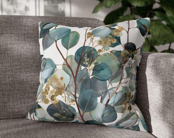 Könnte beinhalten: Quadratisches Zierkissen mit einem botanischen Eukalyptusblatt-Design. Das Kissen hat einen weißen Hintergrund mit teal-, grünen und goldfarbenen Blättern und Zweigen. Das Kissen liegt auf einem grauen Sofa.