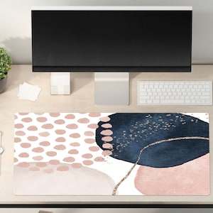 May include: A desk mat with an abstract design featuring pink and blue watercolor shapes with gold glitter accents. The mat is on a light wood desk with a computer monitor, keyboard, mouse, and a potted plant.