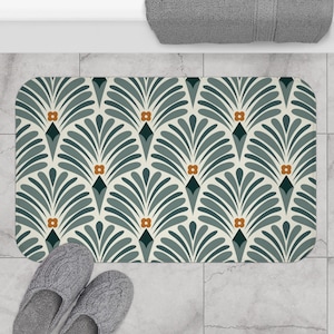 Puede incluir: Una alfombra de baño rectangular con un patrón repetitivo en forma de abanico en tonos turquesa, gris y crema, acentuado con pequeños diamantes naranjas y verde oscuro. Zapatillas grises en la parte inferior izquierda. Una toalla gris en la parte superior derecha.