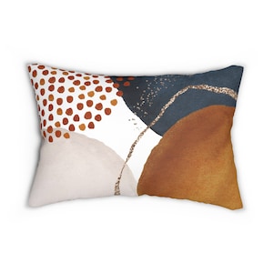 Puede incluir: Una almohada decorativa con un diseño abstracto que presenta formas de acuarela marrones, beige y azules con acentos de purpurina dorada.