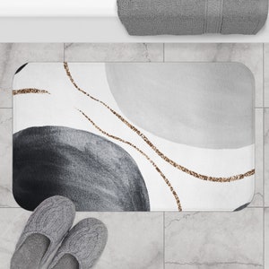 Può includere: Un tappetino da bagno bianco con un design astratto grigio e bianco con accenti di glitter dorati. Il design include due grandi cerchi e due linee sottili.