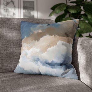 May include: Square decorative pillow with an abstract watercolor design. The pillow features a blend of blue, tan, and white hues, creating a soft, cloud-like pattern. The pillow is placed on a gray sofa, suggesting a home decor theme.