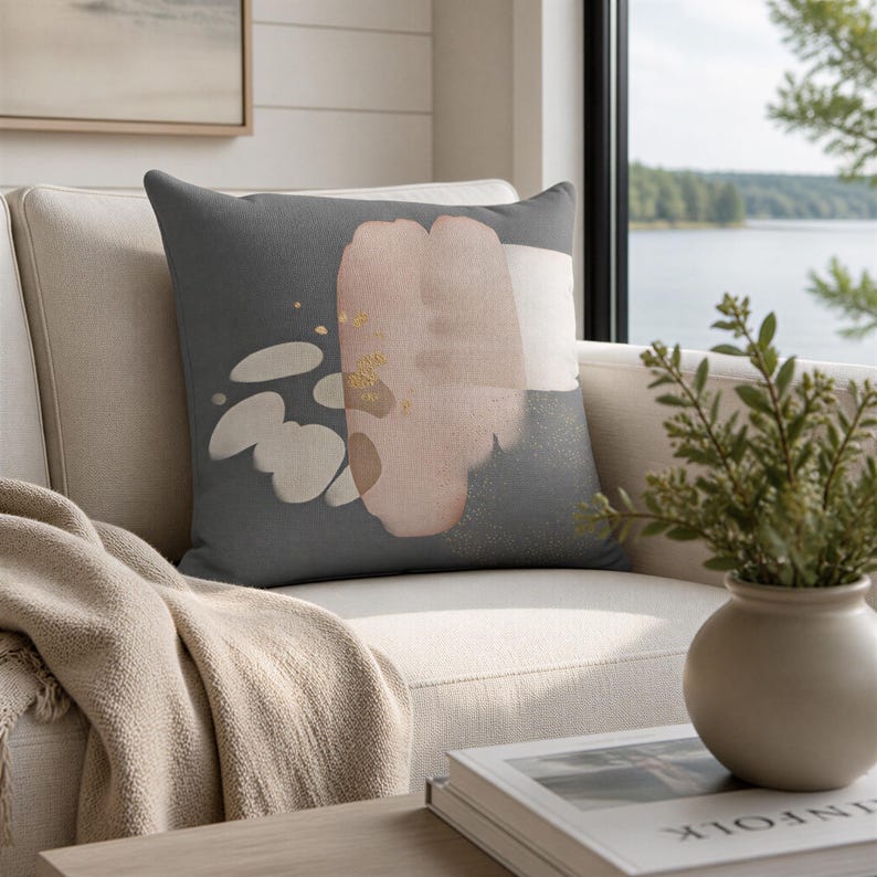 a cozy living room with a gray throw pillow on a beige couch, a white vase with greenery, and a book on a wooden coffee table. The room has a window overlooking a scenic view of a lake and trees.