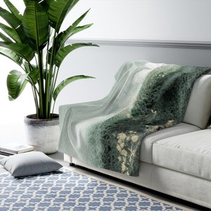 Peut inclure: Une couverture en jetée verte et blanche avec un motif marbré. La couverture est drapé sur un canapé blanc avec une plante verte dans un pot au premier plan.