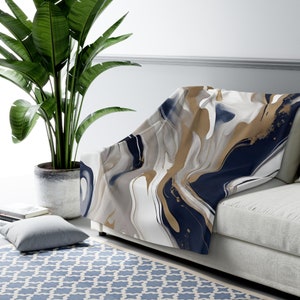 May include: A throw blanket with an abstract design in navy blue, gold, white, and gray. The blanket is draped over a light gray sofa. A large potted plant is to the left of the sofa, and a blue patterned rug is on the floor.