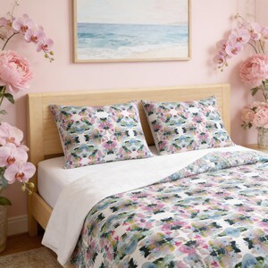 May include: A bedding set featuring two rectangular pillows and a quilt with a white base and a repeating abstract watercolor pattern in shades of pink, blue, and green. The bedding is displayed on a light wood headboard against a pink wall.