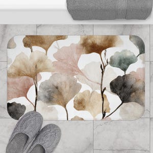 Peut inclure: Tapis de bain rectangulaire avec un motif aquarelle de feuilles de ginkgo dans des tons de marron, rose, gris et vert sur fond blanc. Le tapis est posé sur un sol carrelé, avec des chaussons gris et une serviette grise.
