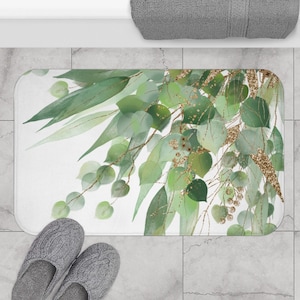 Puede incluir: Una alfombra de baño blanca con un diseño de acuarela verde y dorado de hojas y ramas. El diseño presenta una rama frondosa con hojas verdes y detalles de purpurina dorada.
