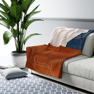 May include: A soft, plush throw blanket with a geometric pattern in shades of rust, navy blue, and white. The blanket is folded and draped over a white couch.
