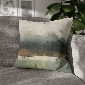May include: A throw pillow with a beige background and an abstract design of blue, green, and brown brushstrokes. The pillow is sitting on a gray couch.