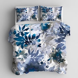 May include: A bedding set featuring a duvet cover and two pillowcases. The set showcases a watercolor floral design in shades of blue, gray, and white. The pattern includes stylized leaves and flowers, creating a soft, artistic aesthetic for the bedroom.