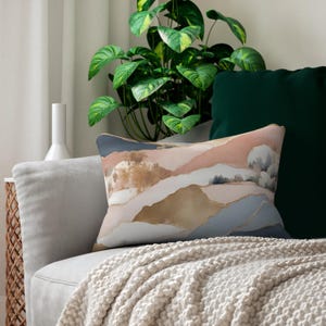 May include: A rectangular decorative pillow with an abstract watercolor design. The pillow features shades of pink, blue, and gold, resembling a landscape. The pillow is placed on a gray sofa with a cream-colored throw blanket.