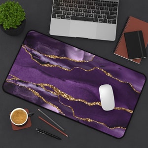 May include: A rectangular desk mat with a purple watercolor design and gold glitter accents. A white computer mouse sits on the mat. A laptop, notebooks, pens, and a small potted plant are also visible.