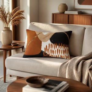 May include: A rectangular throw pillow featuring an abstract design with bold blocks of burnt orange, black, and cream. The pattern includes a textured gold glitter line, a large black circle, and a series of vertical orange brushstroke shapes.