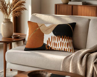 Può includere: Un cuscino rettangolare con un design astratto caratterizzato da blocchi audaci nei colori arancione bruciato, nero e crema. Il motivo include una linea glitterata dorata testurizzata, un grande cerchio nero e forme verticali a pennellata.