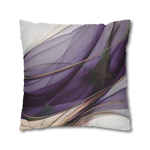 Peut inclure: Coussin carré avec un motif abstrait. Le coussin présente des couches fluides de violet, d'or et de blanc, créant un aspect doux et artistique. Le motif a un effet aquarelle.