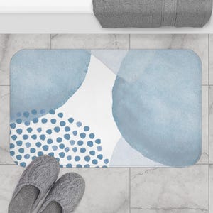 Peut inclure: Un tapis de bain rectangulaire avec un motif aquarelle dans les tons de bleu et de blanc. Le motif présente de grandes formes bleues douces et une section de points bleus. Des pantoufles grises sont au bas du tapis.