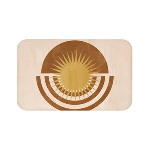 May include: A brown and beige bath mat with a stylized sun design in the center. The sun is yellow with brown rays and is surrounded by two brown concentric circles.