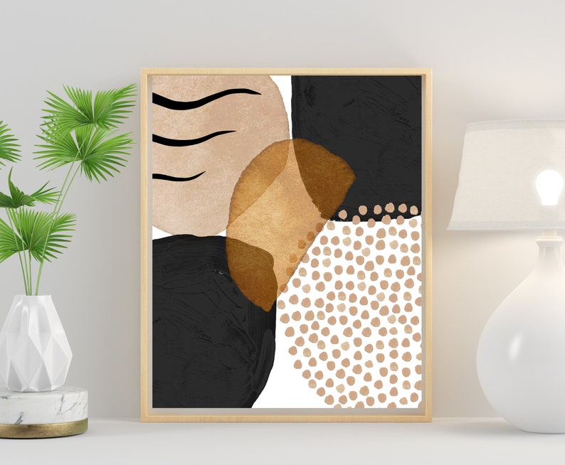 May include: Abstract art print in a wooden frame. The artwork features a composition of black, tan, and gold shapes, including circles, dots, and brushstrokes. The overall design is modern and minimalist, suitable for home decor.