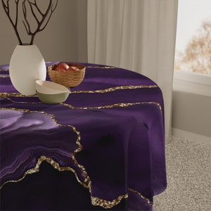 May include: A round purple tablecloth with gold veining, set on a table. A white vase with branches, a small basket of apples, and two bowls are on the table. The tablecloth has a marbled design.