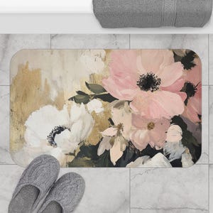 May include: Rectangular bath mat featuring a floral design with pink and white flowers, green leaves, and a gold background. The mat is placed on a marble tile floor, with gray slippers and a gray towel visible.