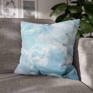 May include: Square throw pillow with a sky-blue and white cloud design. The pillow is placed on a gray sofa. The pillow's design features a realistic depiction of fluffy clouds against a light blue sky, creating a serene aesthetic.